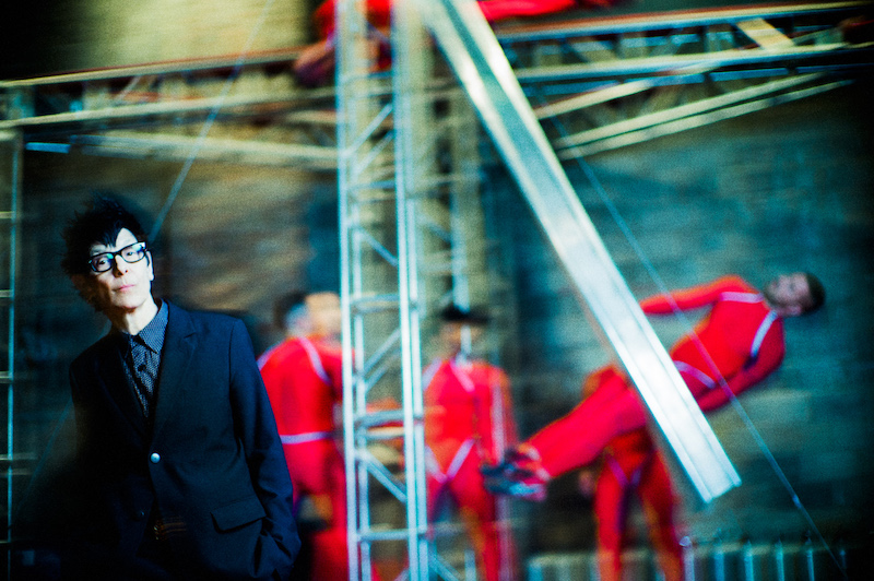 Elizabeth Streb in black acetate glasses and sharp suit. Dancers in red are blurred in the background.